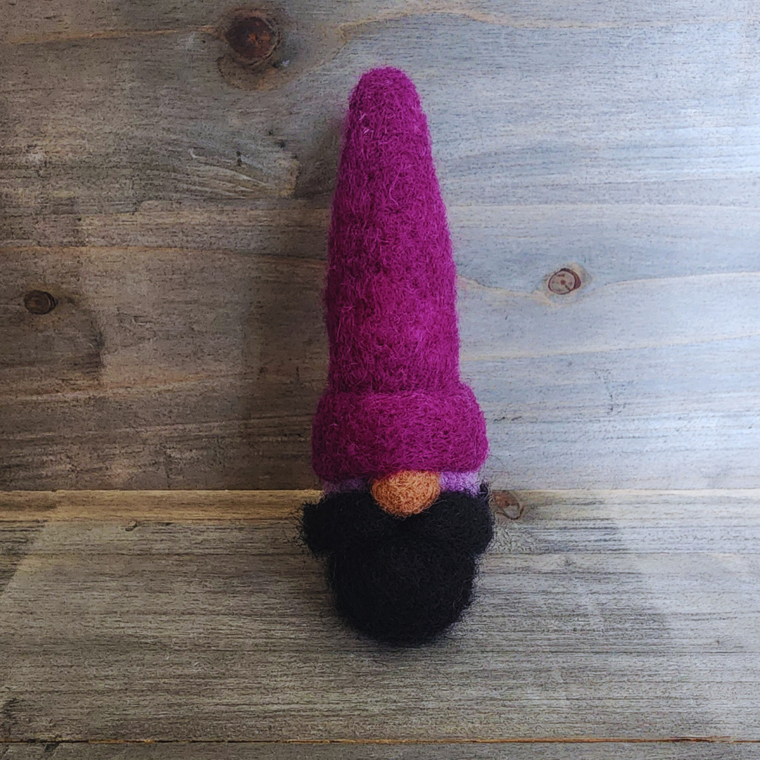 A handmade needle-felted gnome with a deep purple pointy hat, a light-colored nose, and a black beard, with a lavender body partially visible, standing centered against a rustic wooden background.