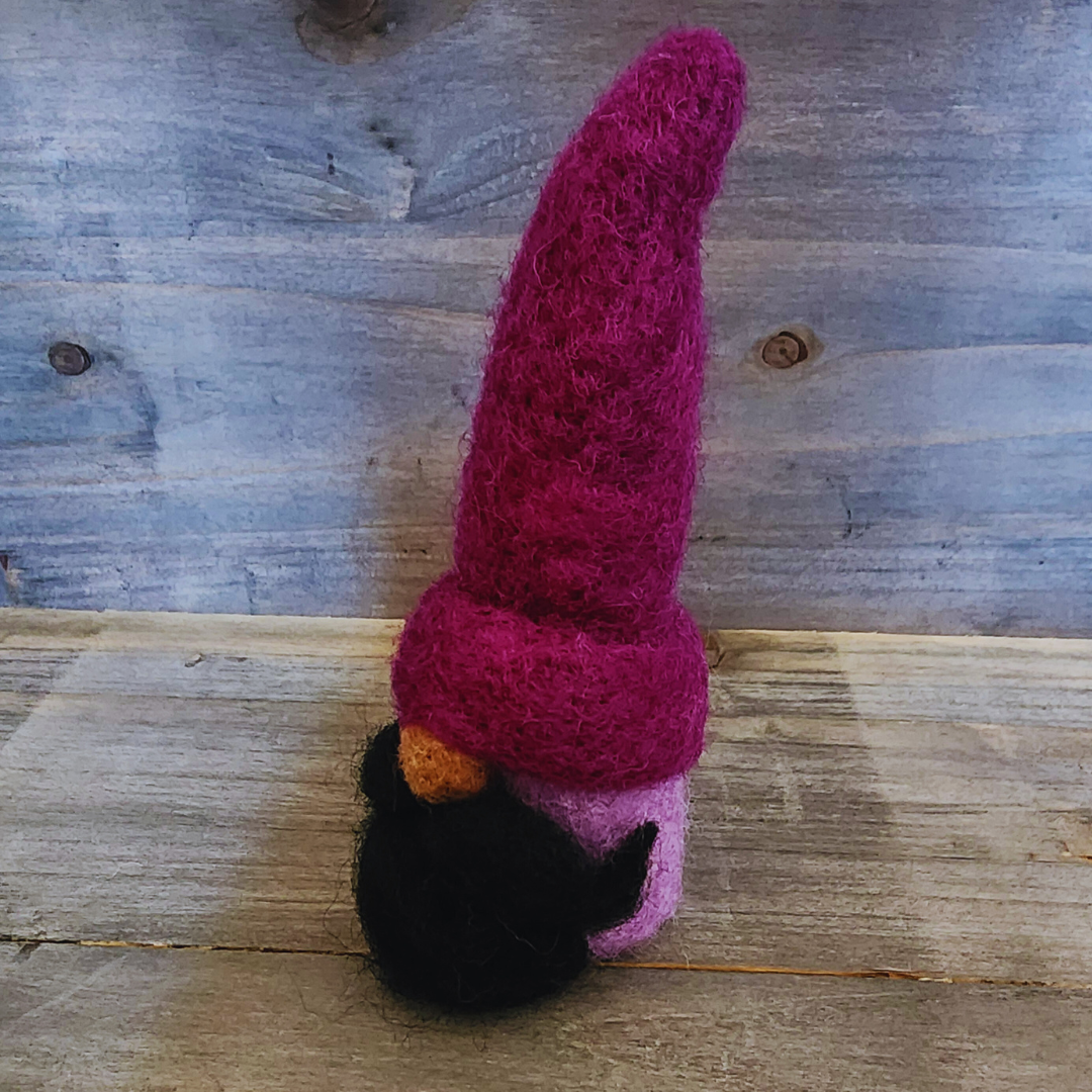 A handmade needle-felted gnome with a deep purple pointy hat and a black beard, viewed from a side angle, showing its lavender body and nose, against a rustic wooden background.