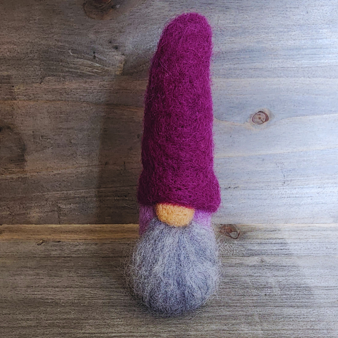 A handmade needle-felted gnome with a deep purple pointy hat, a light-colored nose, and a light gray beard, with a lavender body partially visible, standing centered against a rustic wooden background.
