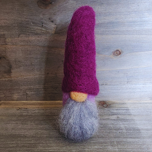A handmade needle-felted gnome with a deep purple pointy hat, a light-colored nose, and a light gray beard, with a lavender body partially visible, standing centered against a rustic wooden background.