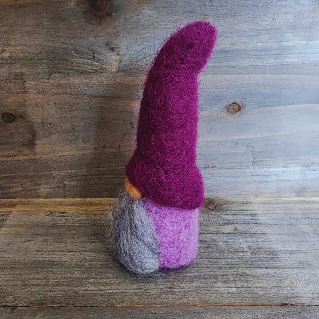 A handmade needle-felted gnome with a deep purple pointy hat, a light-colored nose, and a light gray beard, viewed from a side angle, showing its lavender body, against a rustic wooden background.