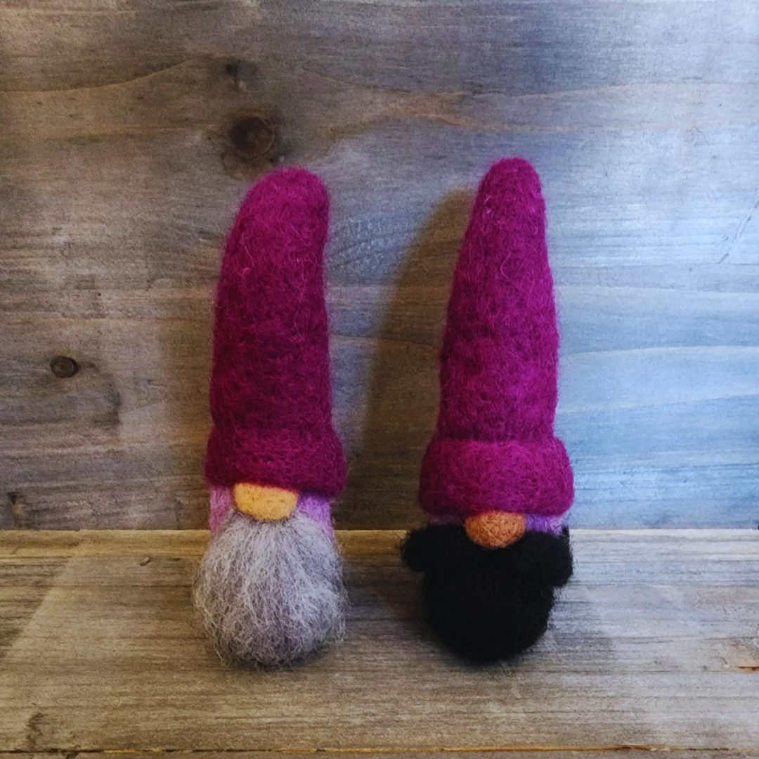 A group of two handmade needle-felted gnomes with deep purple pointy hats, showing one with a light gray beard and the other with a black beard, standing together on a wooden surface.