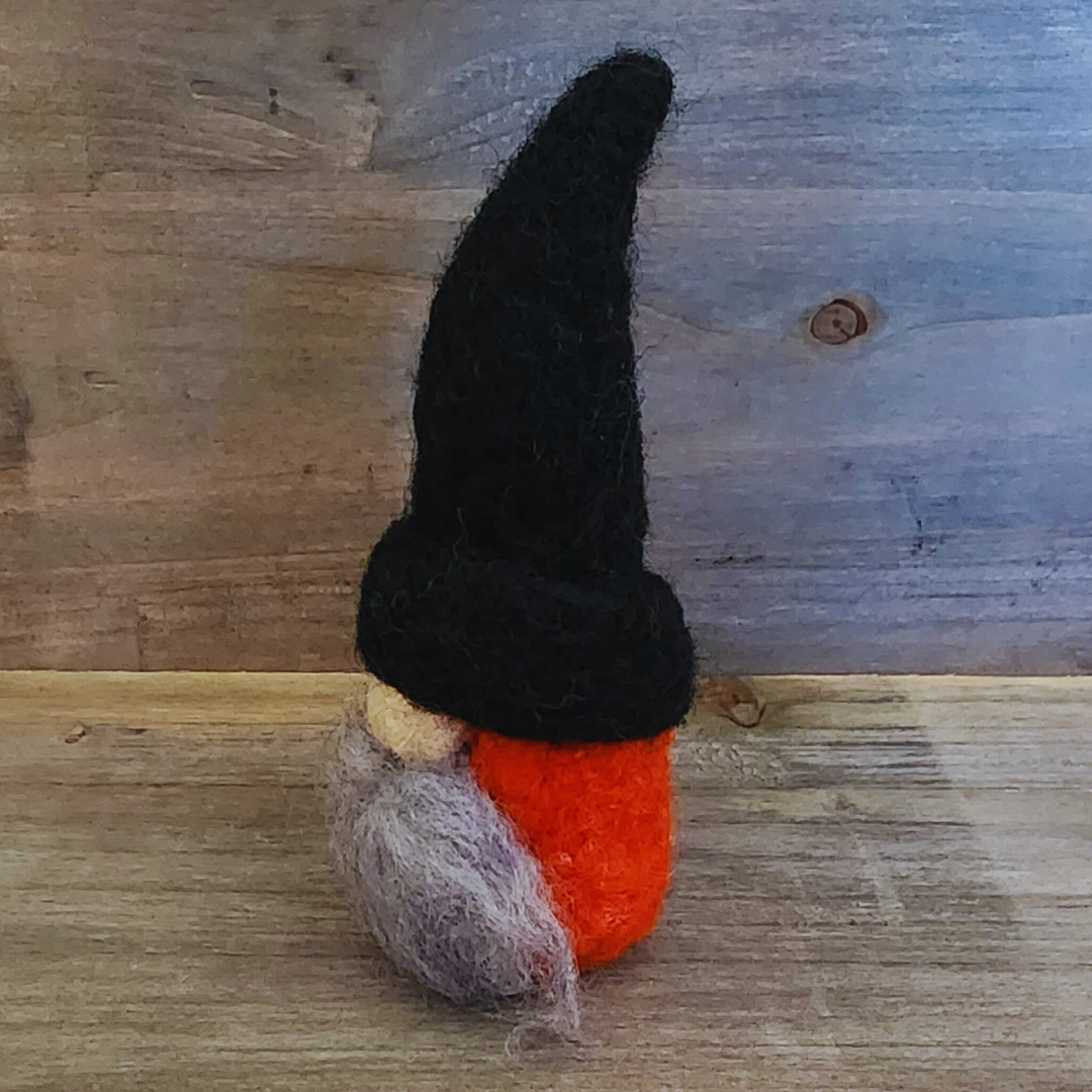 A handmade needle-felted gnome with a black pointed hat and an orange body, viewed from a side angle, showing its light gray beard and nose, against a rustic wooden background.
