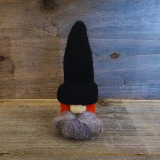 A handmade needle-felted gnome with a black pointed hat, an orange body, a light gray beard, and a light-colored nose, standing centered against a rustic wooden background.