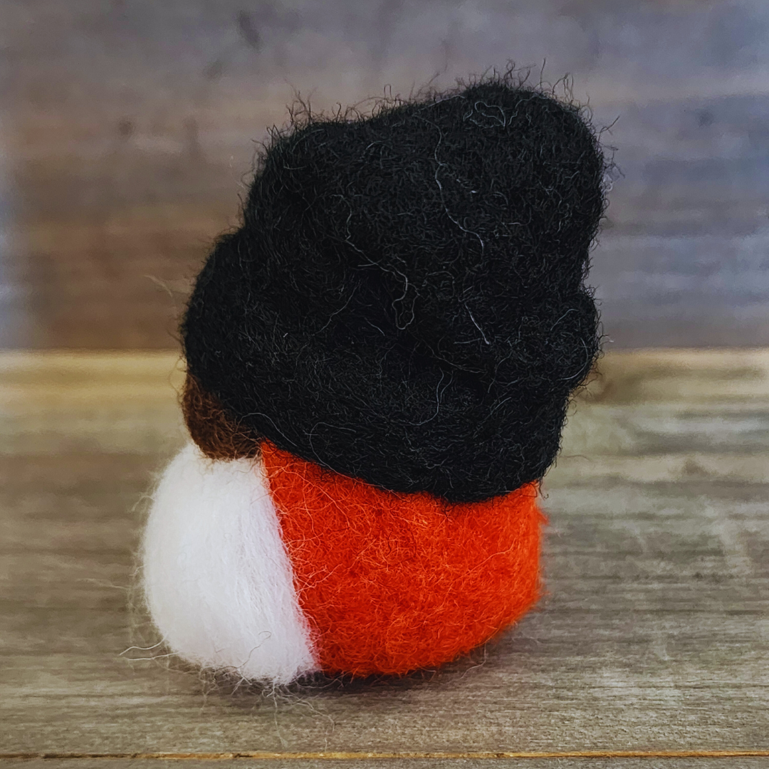 A handmade needle-felted gnome with a black rounded hat, an orange body, a white beard, and a light-colored nose, viewed from a slight angle against a rustic wooden background.