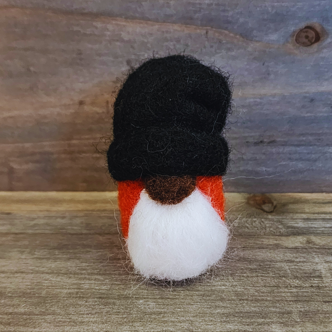 A handmade needle-felted gnome with a black rounded hat, an orange body, a white beard, and a light-colored nose, standing centered against a rustic wooden background.