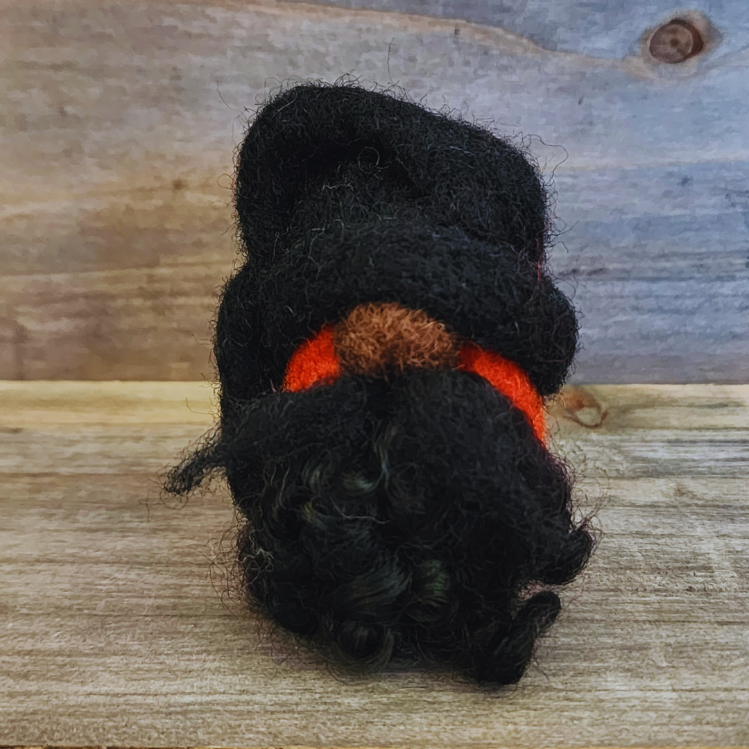 A handmade needle-felted gnome with a black rounded hat and an orange body, viewed from a side angle, showing its curly black beard and nose, against a rustic wooden background.