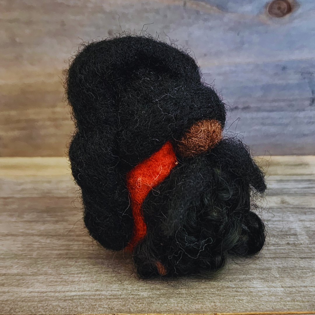 A handmade needle-felted gnome with a black rounded hat and an orange body, viewed from a top-down angle, showing its curly black beard and nose, on a wooden background.