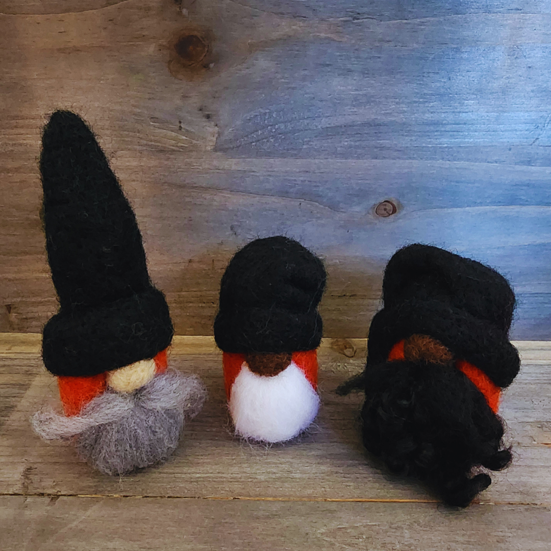 A group of three handmade needle-felted gnomes with orange bodies and black hats, featuring varying beard styles (light gray, white, and curly black), standing on a wooden surface.