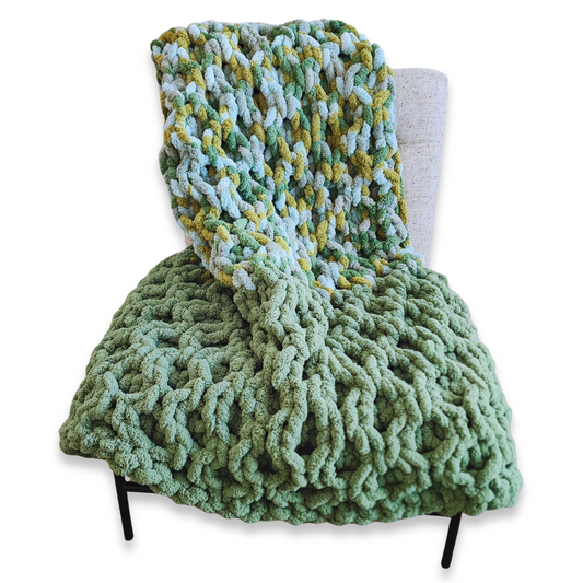 A handmade hand-crocheted blanket with a honeycomb stitch, featuring a solid light green bottom half and a multicolored top half with shades of green, yellow, and light blue, draped over a light-colored chair.