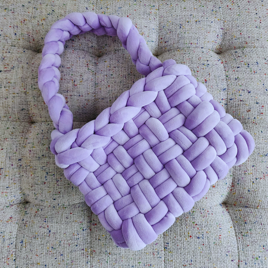 A large handmade basket weave handbag in soft lilac velvet, with a thick braided handle, positioned diagonally on a textured light-colored upholstered surface.