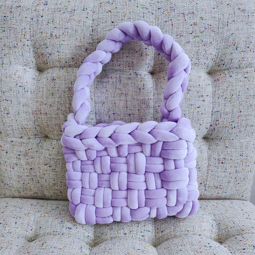 A large handmade basket weave handbag in soft lilac velvet, with a thick braided handle, sitting upright on a textured light-colored upholstered surface.