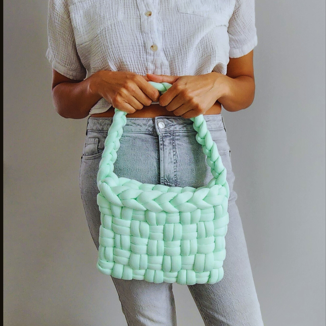 A person wearing a white shirt and light-colored jeans holds a large handmade basket weave handbag in mint green with both hands, positioned in front of their torso.