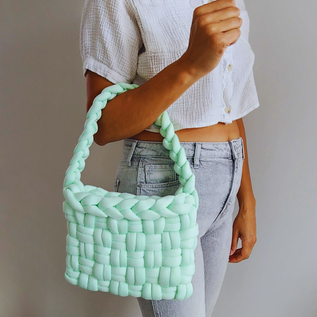 A person wearing a white shirt and light-colored jeans holds a large handmade basket weave handbag in mint green over their shoulder, showing the braided strap and woven texture.