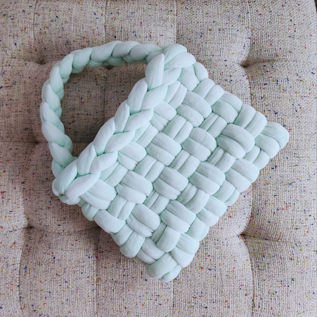 A large handmade basket weave handbag in light mint green velvet, with a thick braided handle, positioned diagonally on a textured light-colored upholstered surface.