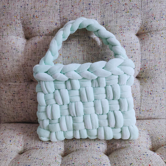 A large handmade basket weave handbag in light mint green velvet, with a thick braided handle, sitting upright on a textured light-colored upholstered surface.