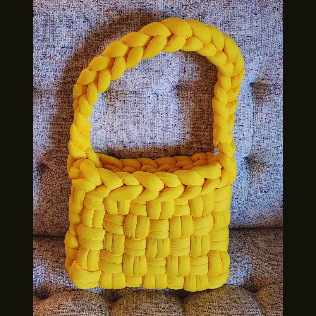 A large handmade basket weave handbag in vibrant yellow, with a long, thick braided handle, sitting upright on a multicolored speckled fabric surface.