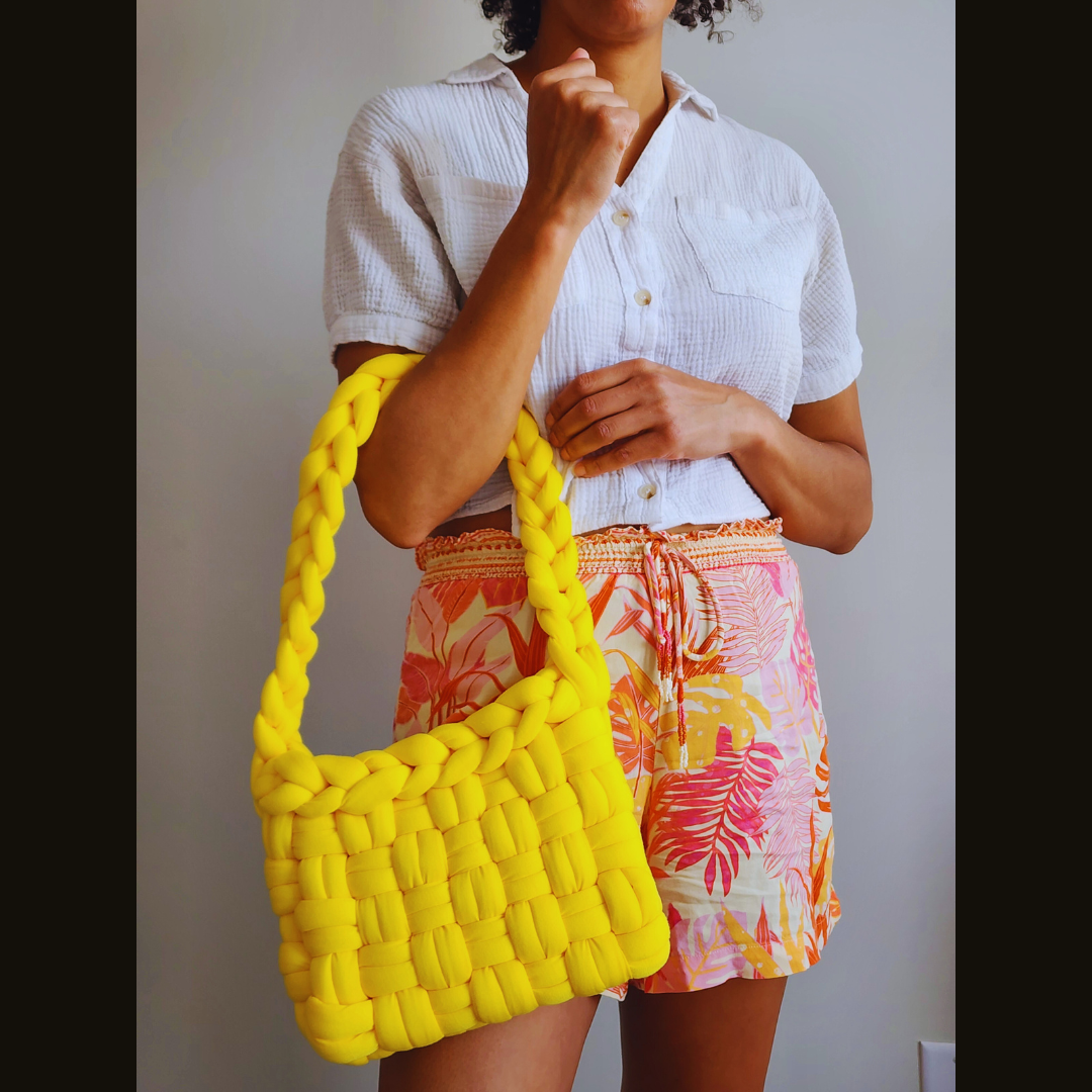 A person wearing a white shirt and tropical print shorts holds a large handmade yellow basket weave shoulder bag over their shoulder, posed against a white wall.
