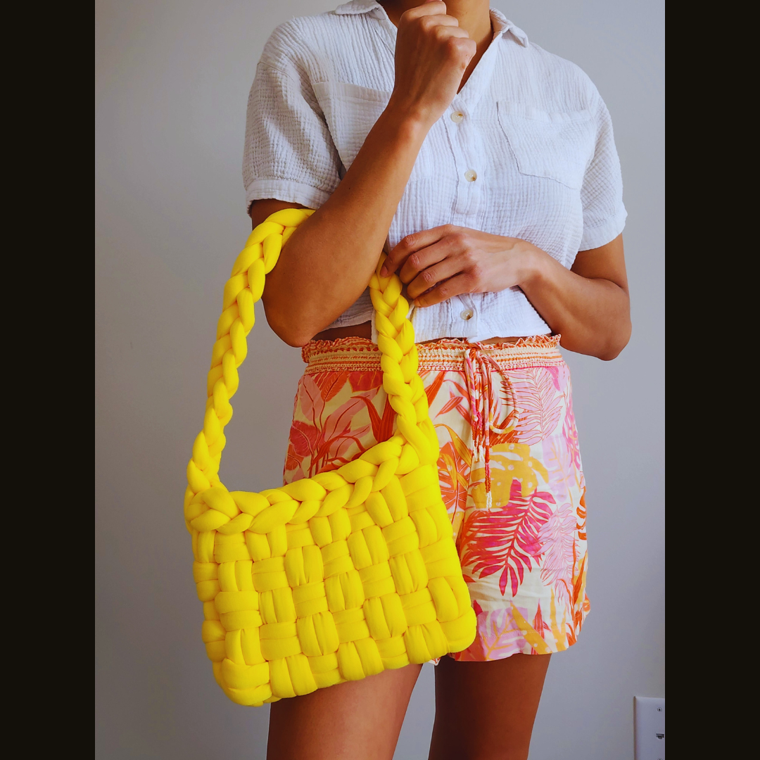 A person wearing a white shirt and tropical print shorts holds a large handmade yellow basket weave shoulder bag, resting it on their arm and hip, posed against a white wall.