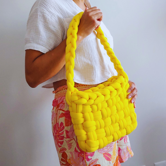 A person wearing a white shirt and tropical print shorts holds a large handmade yellow basket weave shoulder bag with a long braided strap, posed against a white wall.