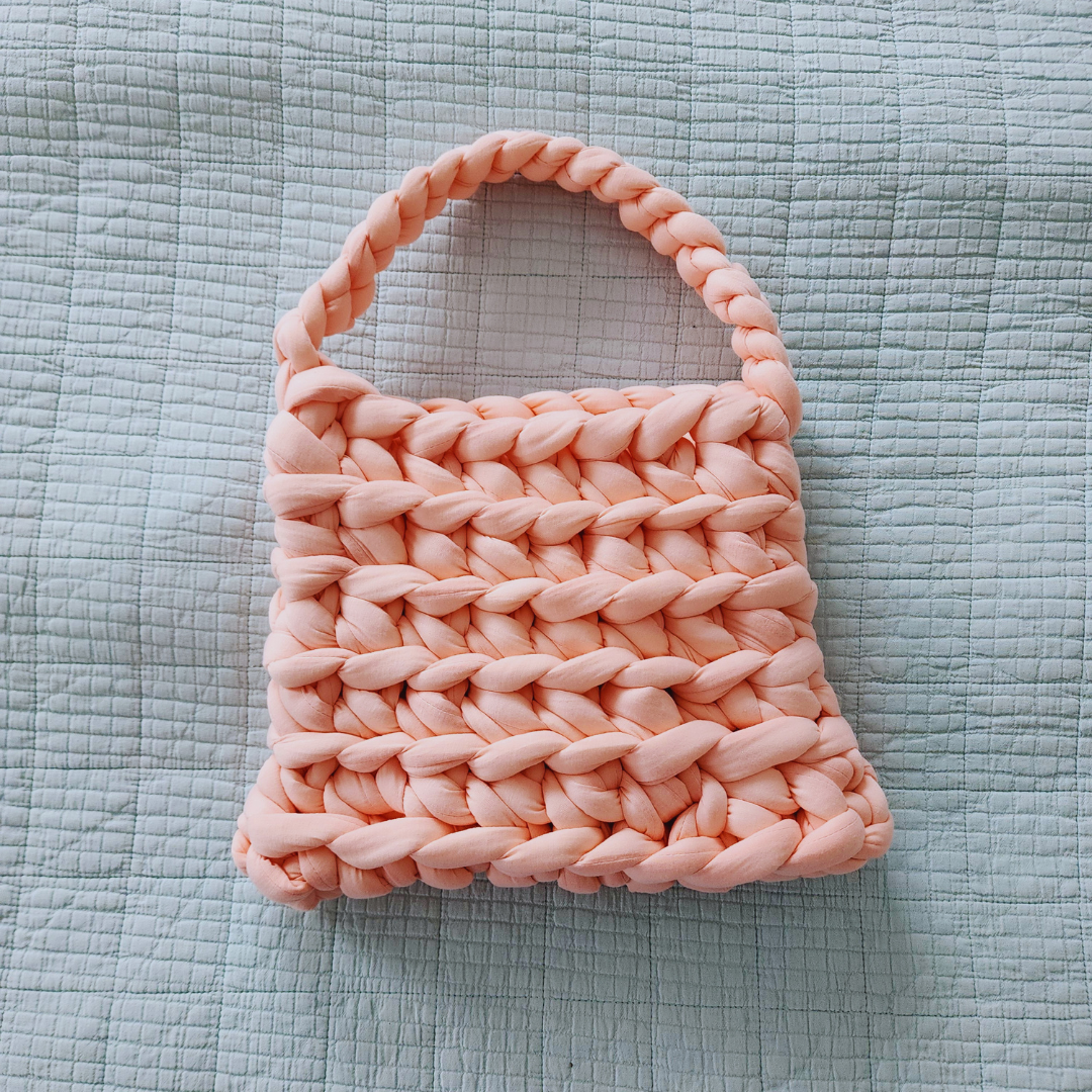 A large handmade ribbed shoulder bag in light coral pink knit yarn, with a thick braided handle, lying flat on a light blue quilted surface.