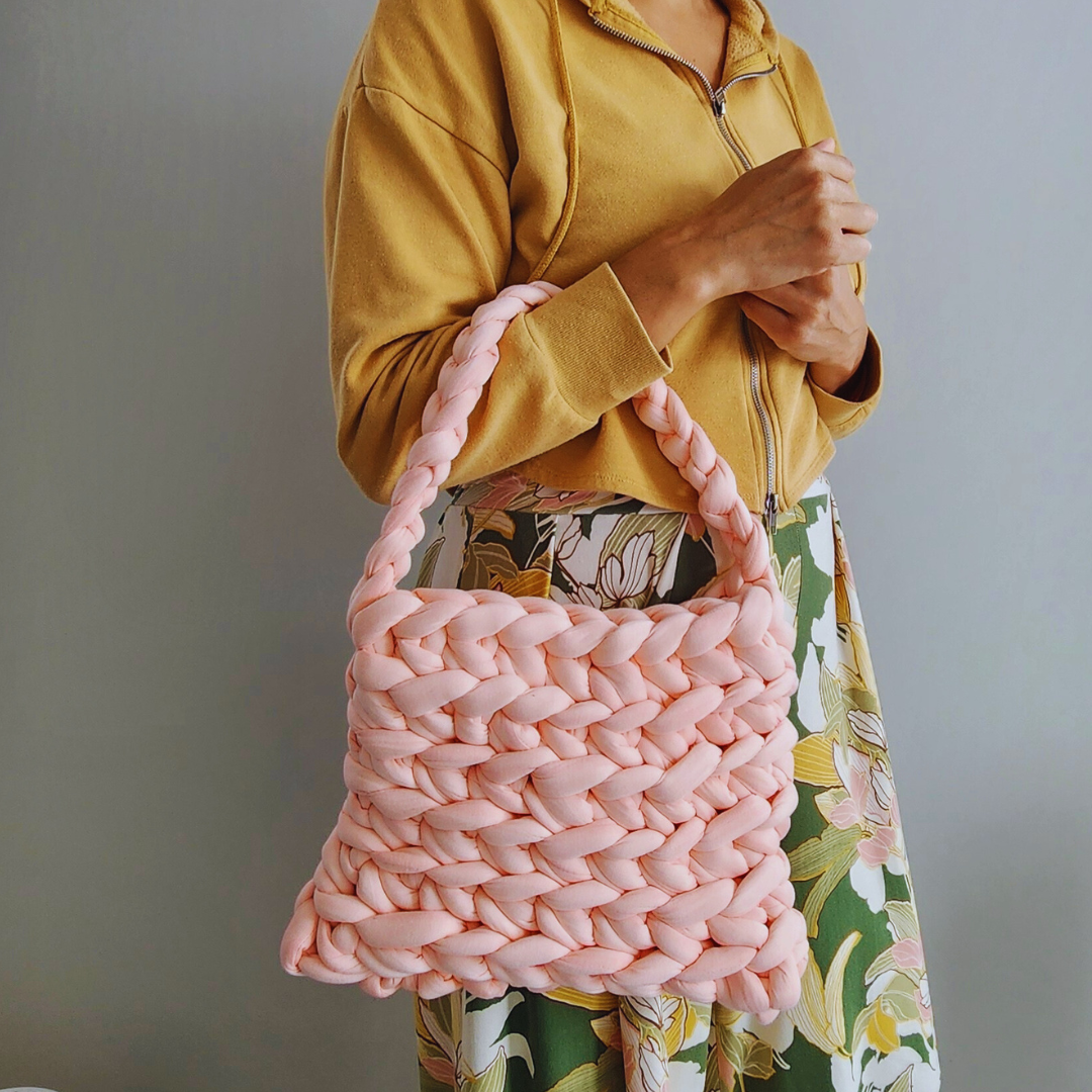 A person wearing a yellow hoodie and a floral skirt holds a large handmade ribbed shoulder bag in light coral pink knit yarn on their arm, showing its woven texture.