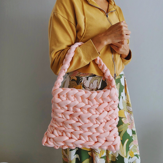 A person wearing a yellow hoodie and a floral skirt holds a large handmade ribbed shoulder bag in light coral pink knit yarn on their arm, showing its woven texture.