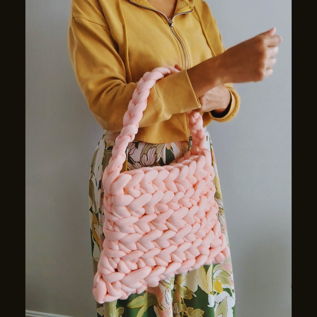 A person wearing a yellow hoodie and a floral skirt holds a large handmade ribbed shoulder bag in light coral pink knit yarn, posed in front of a gray wall.