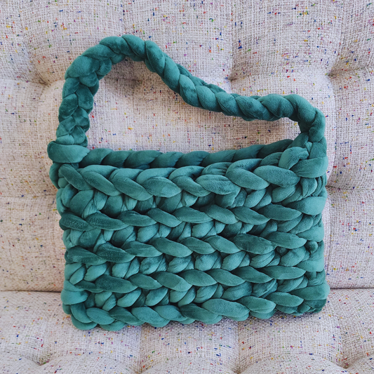 A large handmade ribbed shoulder bag in rich forest green velvet, with a thick braided handle, sitting upright on a multicolored speckled fabric surface.