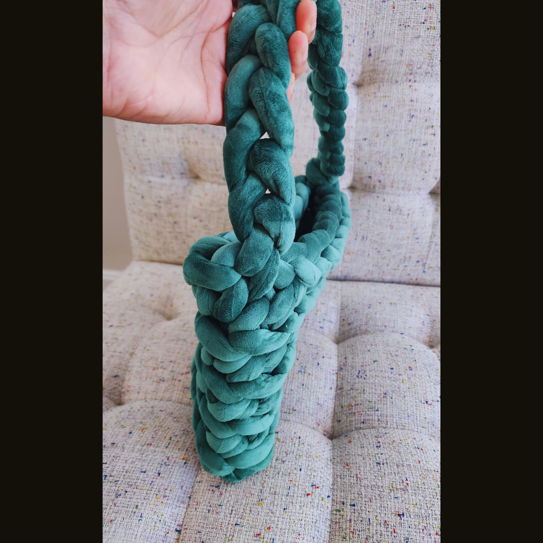 A close-up side view of a large handmade ribbed shoulder bag in forest green velvet, showing the texture of the braided handle and the bag, held by a hand.