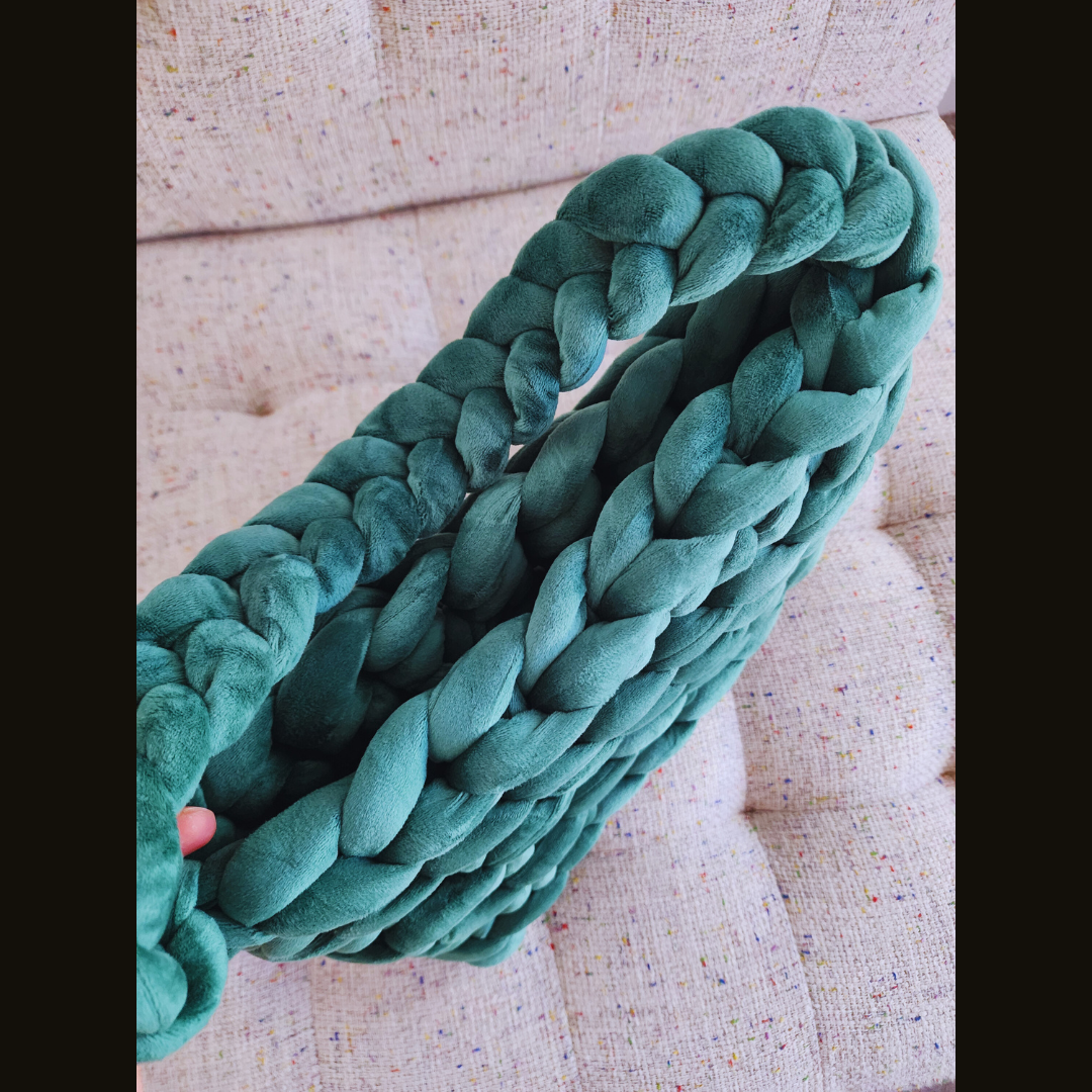 A large handmade ribbed shoulder bag in rich forest green velvet, with a thick braided handle, positioned diagonally on a multicolored speckled fabric surface.