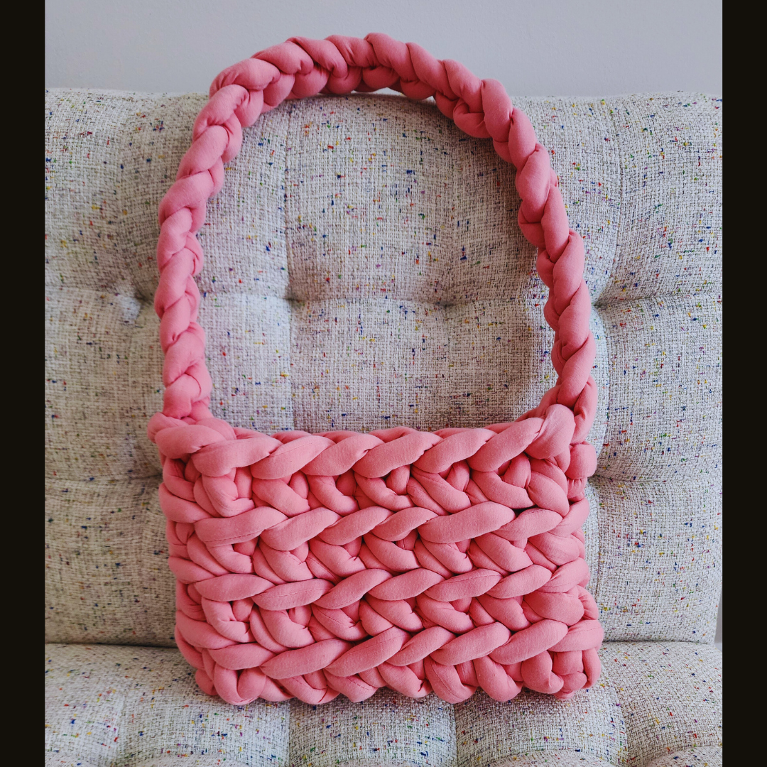 A large handmade ribbed shoulder bag in solid rose pink knit yarn, with a thick braided handle, sitting upright on a multicolored speckled fabric surface.