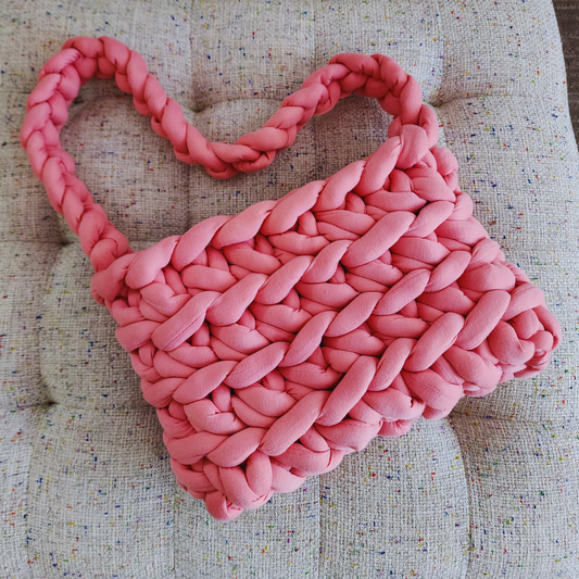 A large handmade ribbed shoulder bag in solid rose pink knit yarn, with its thick braided handle forming a heart shape above the bag, resting on a multicolored speckled fabric surface.