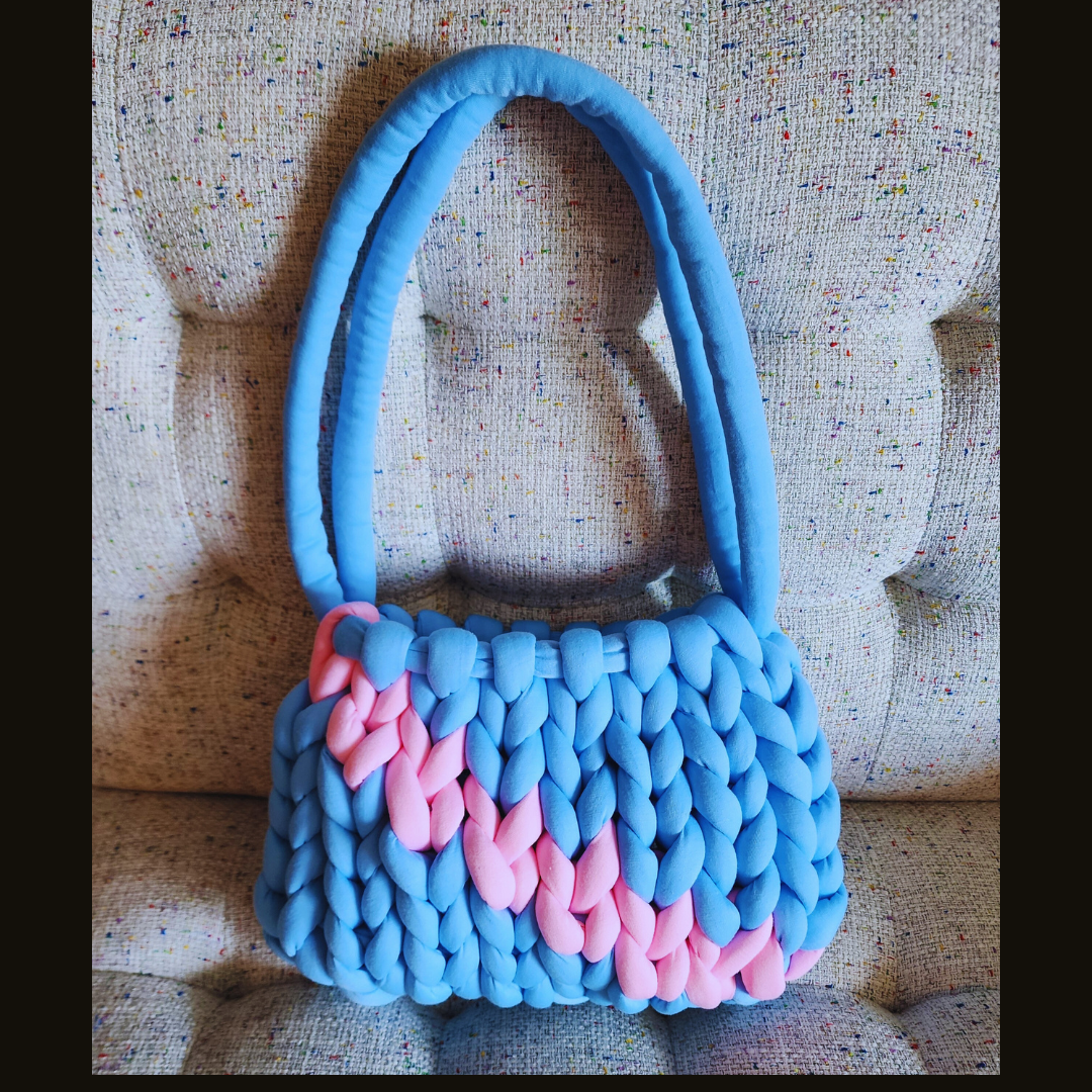 A large handmade single stitch knit handbag in light blue with a vibrant pink chevron design across the front, and blue double straps, sitting upright on a textured upholstered surface.