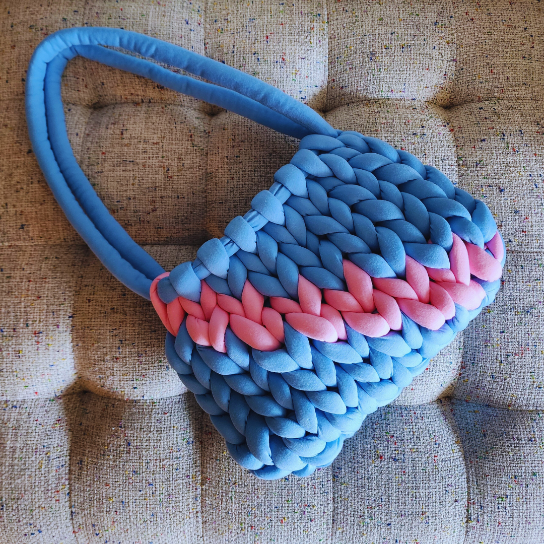 A large handmade single stitch knit handbag in light blue with a vibrant pink horizontal stripe, and blue double straps, positioned diagonally on a textured upholstered surface.