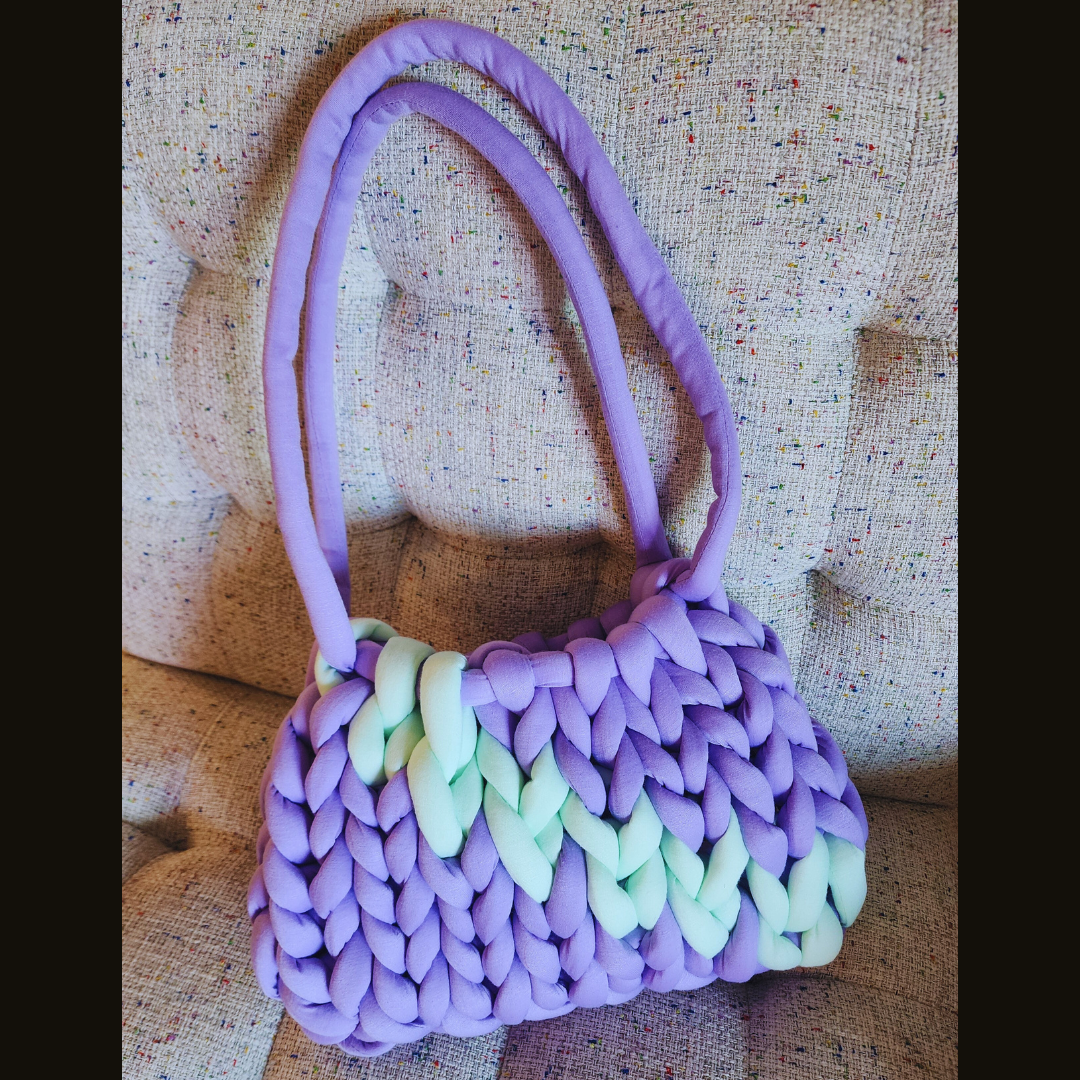A large handmade single stitch knit bag in light purple with a light mint green chevron design across the front, and purple double straps, sitting upright on a textured upholstered surface.
