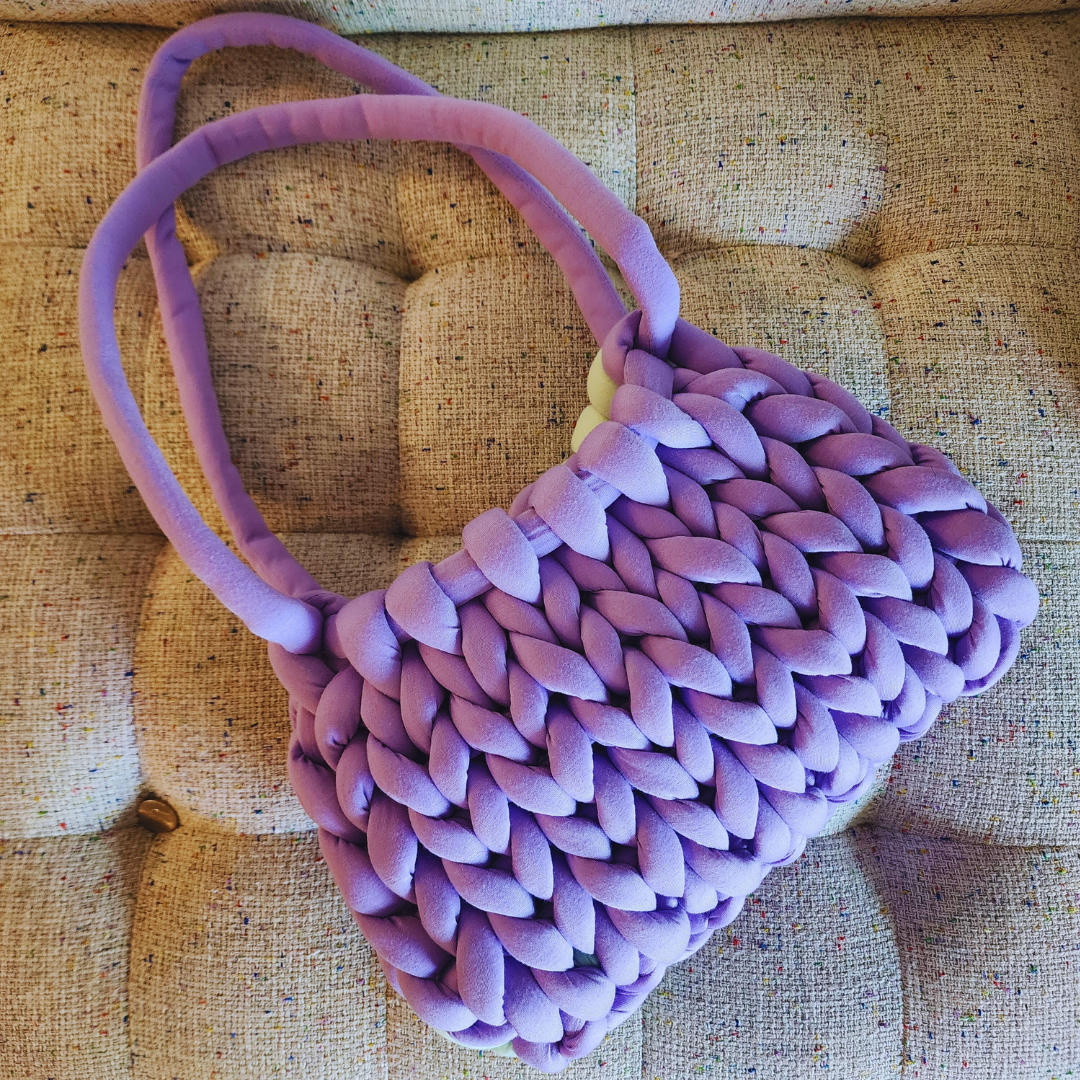 A large handmade single stitch knit bag in solid light purple with matching double straps, sitting upright on a textured upholstered surface.