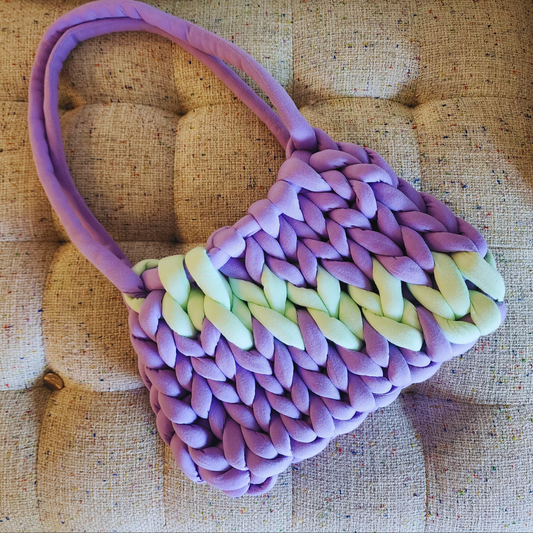 A large handmade single stitch knit bag in light purple with a central horizontal stripe of light mint green, and purple double straps, positioned diagonally on a textured upholstered surface.