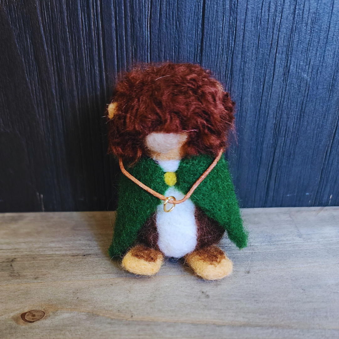 Full-body shot of a needle-felted Frodo Baggins, showing his brown curly hair, green cloak, and the One Ring necklace.