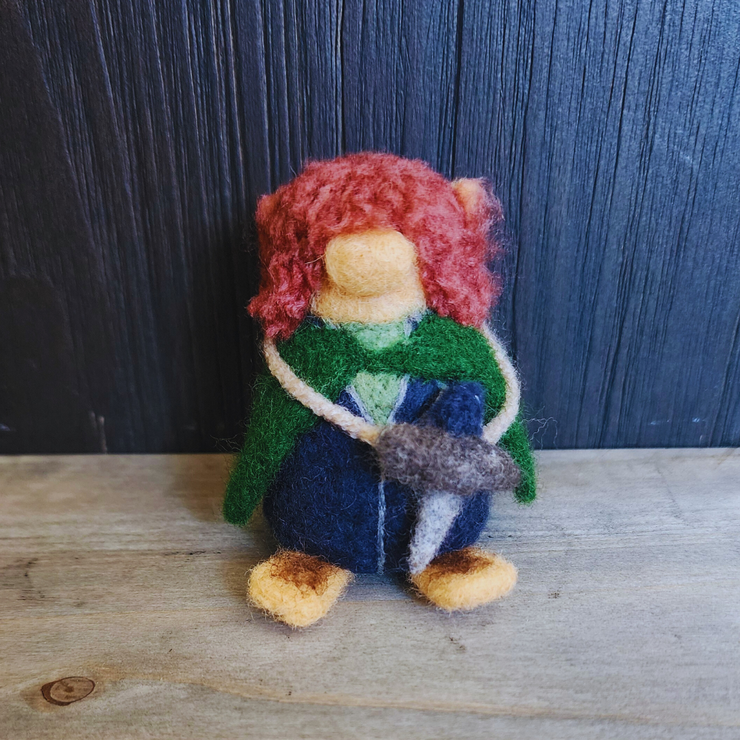Full-body shot of a needle-felted Pippin Took figure, with reddish-brown curly hair, wearing a blue and green outfit with a small sword slung across his chest.