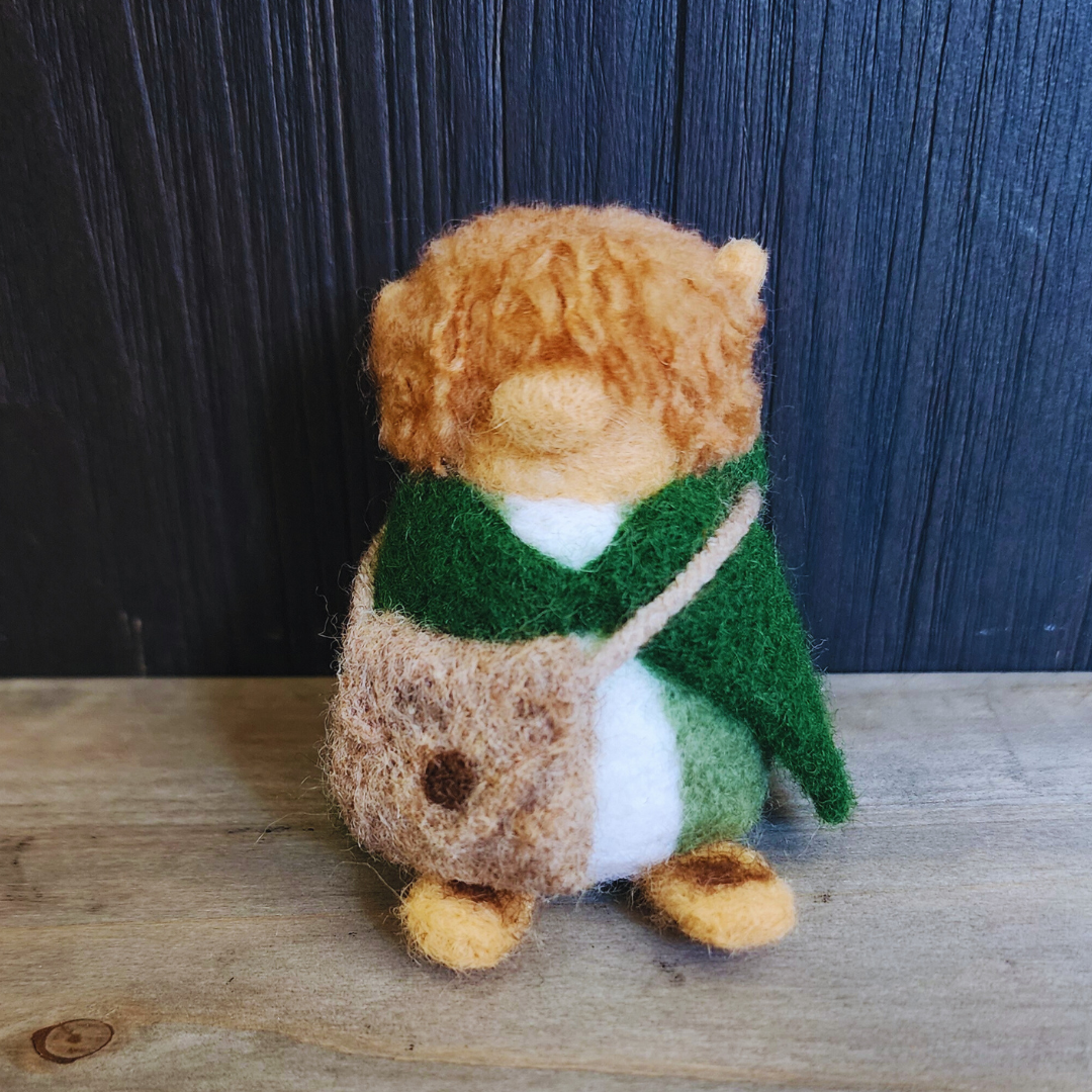 Full-body shot of a needle-felted Samwise Gamgee figure, with tousled reddish-brown hair, wearing a green cloak and carrying a fuzzy brown shoulder satchel.