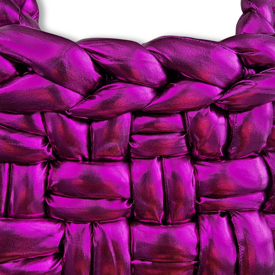 A close-up view of the side and top edge of a handmade chunky knit handbag in shimmering metallic fuchsia, showing its basket weave texture and braided top.