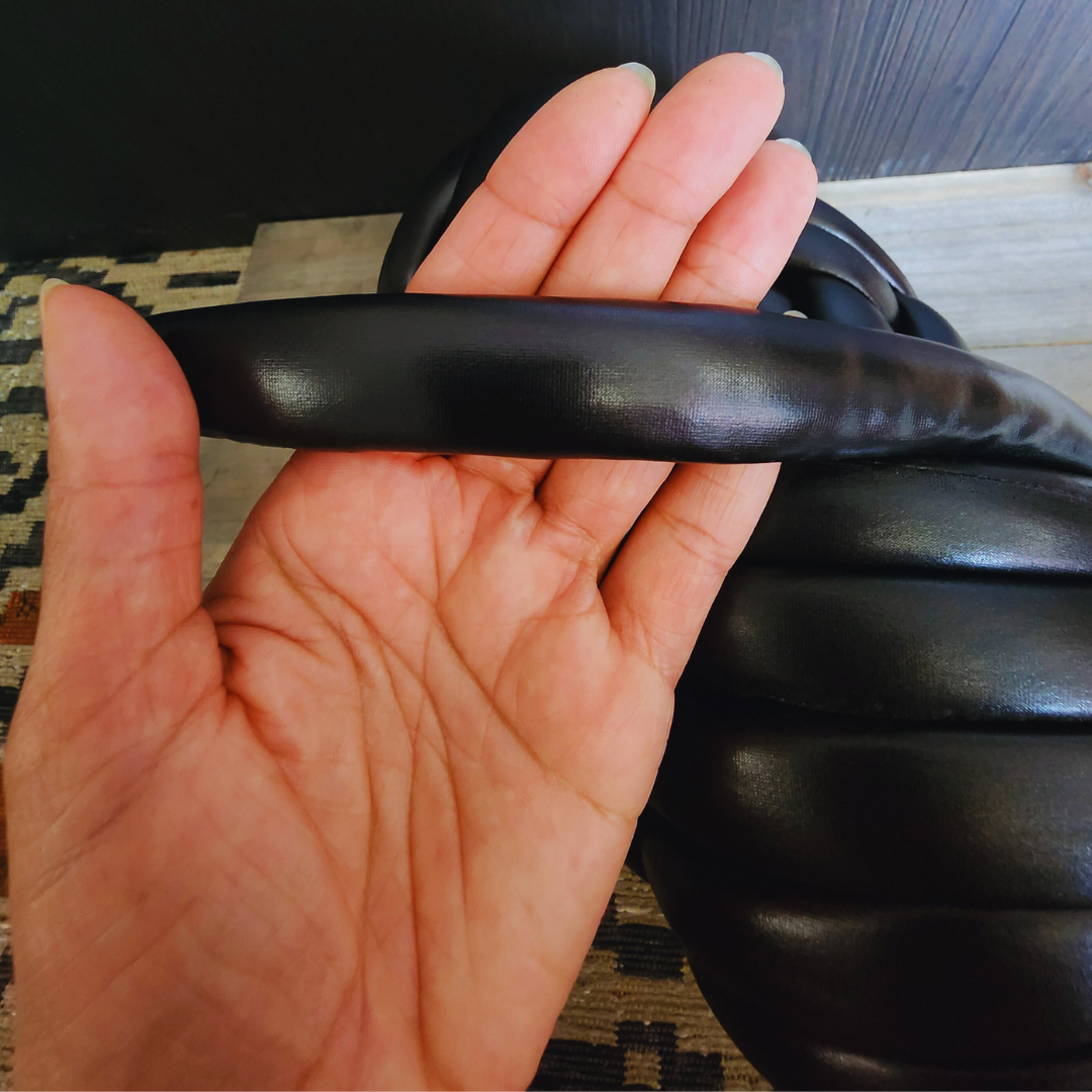 A close-up view of a hand holding a strand of thick, black metallic chunky yarn, showing its width and texture against a dark background.