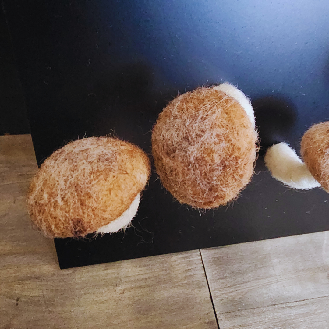 A close-up of two handmade felted brown mushroom caps, showing their textured surface, with white stems partially visible, on a black background against a wooden floor.