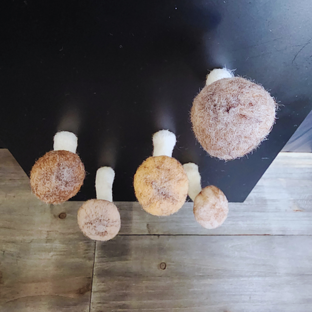 Four handmade felted brown mushroom magnets with white bendable stems, of various sizes and shades, lying unattached on a wooden surface.