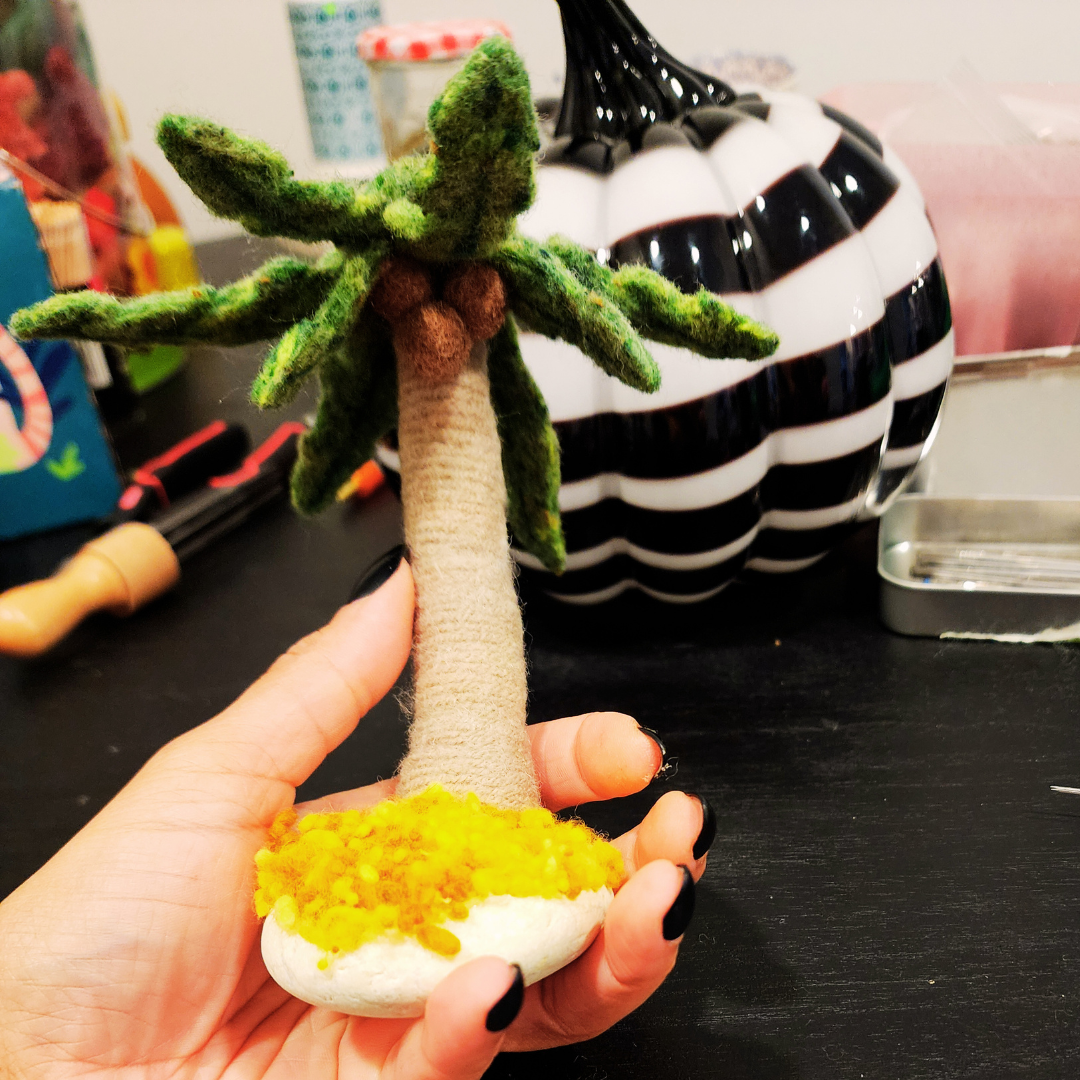 Close-up of a needle-felted miniature palm tree, featuring green fronds and brown coconuts, attached to a white stone base with yellow wool nepps (sand). The tree is being held by a hand with black fingernails.