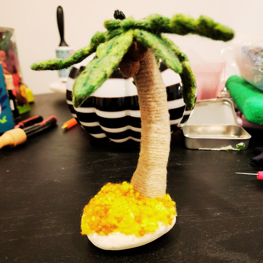 A finished needle-felted palm tree desk ornament, showing its curved trunk, green fronds, and wool nepp sand anchored to a smooth white granite stone.