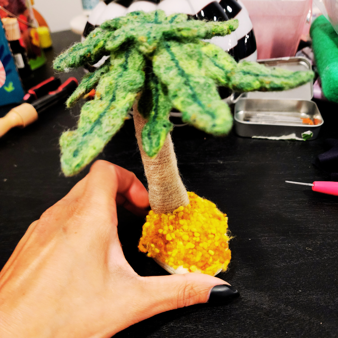 A closer view of the felted palm tree's canopy, showing the textured green wool of the fronds and the coconuts. A hand holds the piece on a dark surface.