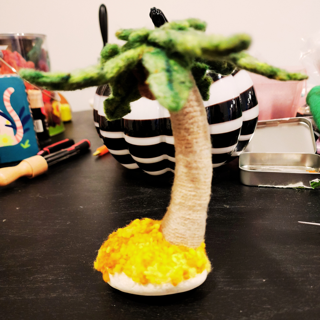 A finished needle-felted palm tree desk ornament, showing its curved trunk, green fronds, and wool nepp sand anchored to a smooth white granite stone.