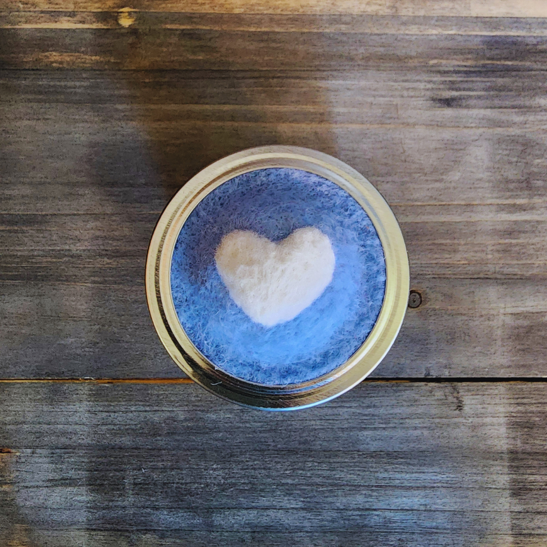 A top-down, centered view of a 4oz quilted mason jar lid. It features a soft blue needle-felted wool base with a white 3D heart felted in the center, held in place by a silver metal ring.